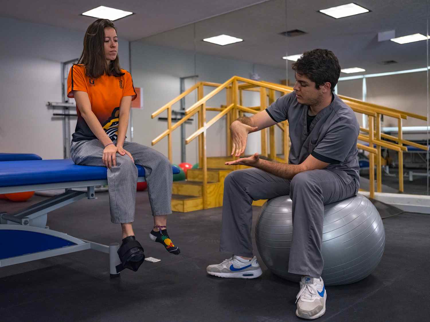 Terapeuta físico sentado en pelota de ejercicio explica técnica de rehabilitación a paciente con férula en el pie.